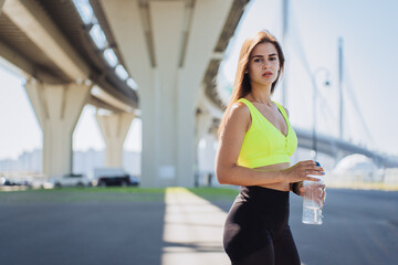 Determined woman in neon workout attire holding a water bottle, standing under a bridge on a sunny...