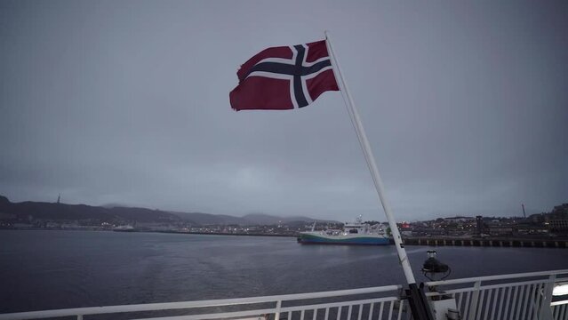 waving norwegian flag on flagpole on deck of sailing ferry with city of bodo on background in slow motion in norway