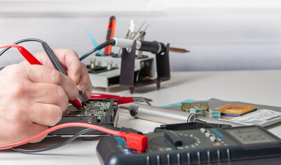Close-up of a technician's hands in a workshop. The repairer is using a multimeter to measure the parameters of an electronic device on a table.