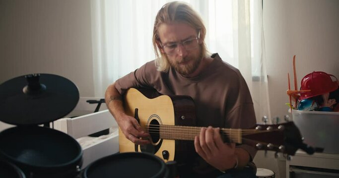 A blond man in glasses with a beard in a brown T-shirt plays a yellow acoustic guitar near him there is a drum set development of talents and hobbies at home