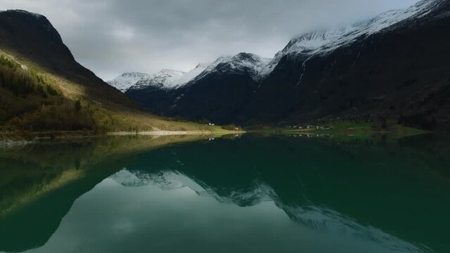 aerial cinematic movement towards valley with houses in sun over Oldevatnet lake with mountains reflected in blue water in norway