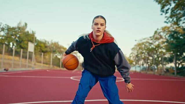 Close up a blonde girl in a sports uniform runs and maneuvers with an orange sword and throws the ball into the hoop during her game of basketball on the street court