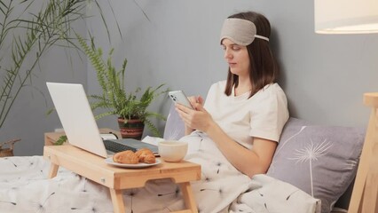 Brown haired woman with sleeping mask on head sitting in bed in front of laptop display using smartphone scrolling online starting remote work during weekend morning