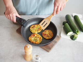 Vegetable pancakes in frying pan held by woman's hands with wooden spatula on the kitchen table....