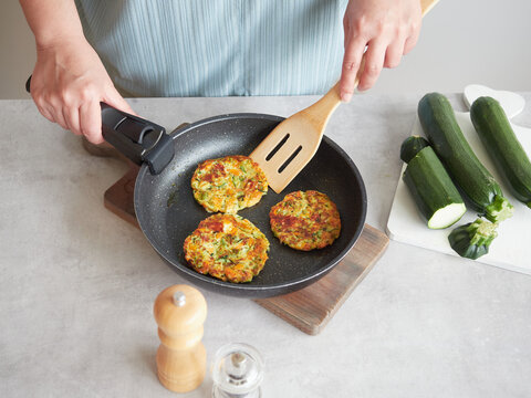 Vegetable pancakes in frying pan held by woman's hands with wooden spatula on the kitchen table. Healthy meal.