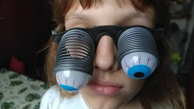 A 9-year-old caucasian girl wearing glasses with eyeballs falling out on springs. A child is preparing for Halloween and trying on funny, scary accessories. Masquerade decoration. Black humor