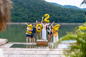 Group of Young Asian man and woman friends celebration happy new year party holding golden number 2024 balloon. People enjoy and fun travel nature mountain together at lake house on holiday vacation.