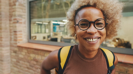 Close-up of young woman in glasses, wearing brown top, with backpack on her shoulders, smiling and...