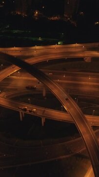 Illuminated complex road junction and cars traffic at night. Drone is flying back. Aerial view. Vertical Video