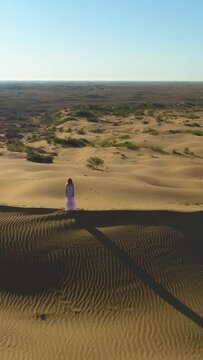 Drone is flying over young lonely woman with long hairs in white dress staying on the top of sand beige dune in desert. Aerial view. Vertical Video