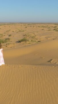 Young lonely woman with long hairs in white dress is staying on sand beige dune in desert. Aerial view. Vertical Video