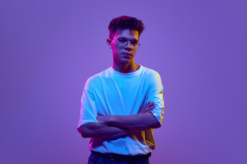 Young buy in casual clothes standing with serious concentrated face and crossed arms against purple...
