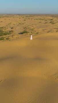 Young lonely woman with long hairs in white dress is staying on the top of sand beige dune in desert. Drone is distancing. Aerial view. Vertical Video