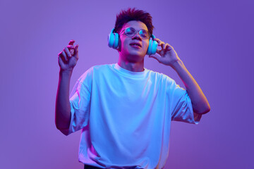 Young guy in white t-shirt and glasses listening to music in headphones against purple background...