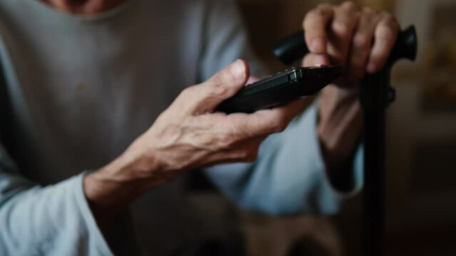 a pensioner with a walking stick sits and presses buttons on the TV remote control. an elderly man spends time at home watching his favorite TV programs