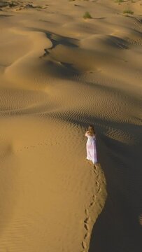 Young lonely woman with long hairs in white dress is walking on a top of sandy beige dune in desert. Aerial view. Camera is tilting down. Vertical Video