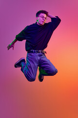 Young man in casual stylish clothes and eyeglasses jumping with positive face against gradient pink background in neon light. Concept of human emotions, youth, delightful, excitement