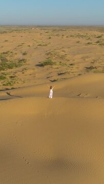 Young lonely woman with long hairs in white dress is staying with raised hands on sand beige dune in desert. Aerial view. Camera is tilting down. Vertical Video