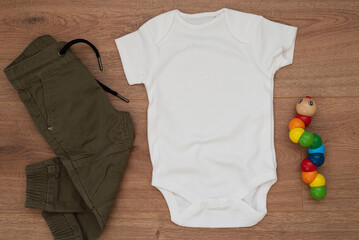 Mockup of white baby bodysuit on wood background with pants and colorful toy. Blank baby clothes template mock up, flat lay styled stock photo.