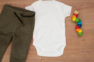Mockup of white baby bodysuit on wood background with pants and colorful toy. Blank baby clothes...