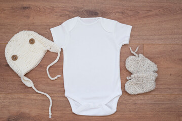 Mockup of white baby bodysuit on wood background with knitted booties and hat. Blank baby clothes template mock up.