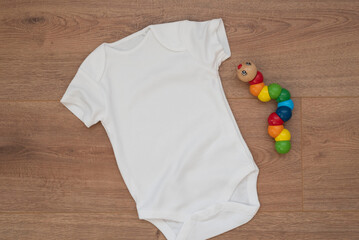 Mockup of white baby bodysuit on wood background with colorful toy. Blank baby clothes template mock up, flat lay styled stock photo