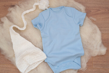 Mockup of blue baby bodysuit on wood background with knitted hat. Blank baby clothes template mock up. Flat lay styled stock photo.