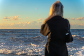 Blonde young woman in a black coat looks at a stormy sea or ocean at sunset, a selective focus....
