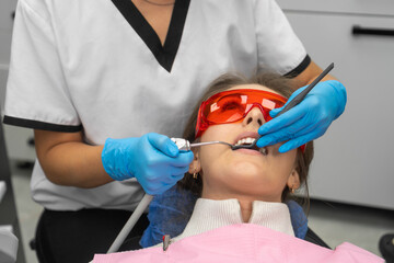 Woman dentist washes the patient's teeth during an oral examination. Young woman wearing safety glasses in a dental chair at a dentist appointment. - obrazy, fototapety, plakaty