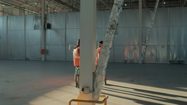 Full length arc shot of three male construction workers with ladder and tool boxes standing in centre of large empty building and speaking during workday