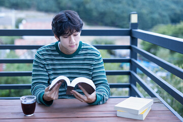 A young Asian Korean university student who is enjoying a rest and leisure life by reading books and drinking coffee in a green natural space