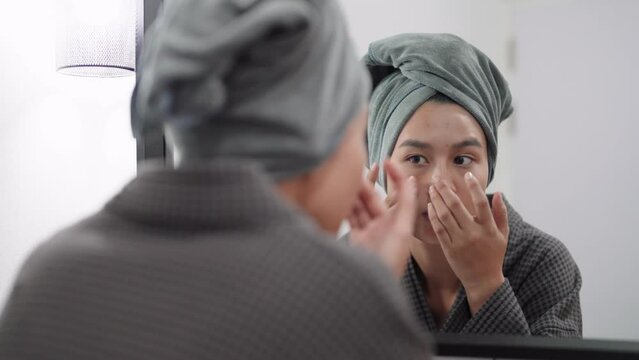 Asian woman is examining her face for blemishes and acne in front of a bathroom mirror, preparing for a shower. Capture skincare routine and self-care moments for a radiant image