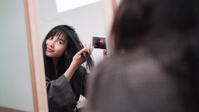 An Asian woman blow-drying her hair in front of a large mirror after washing and styling it at home. Capture the essence of beauty and self-care.