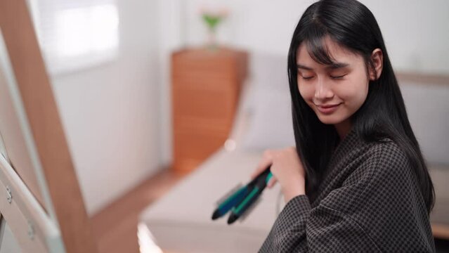 Asian woman using a hair straightener to achieve smooth hair in front of a large mirror after washing and styling at home. Showcase the beauty routine for enhanced sales