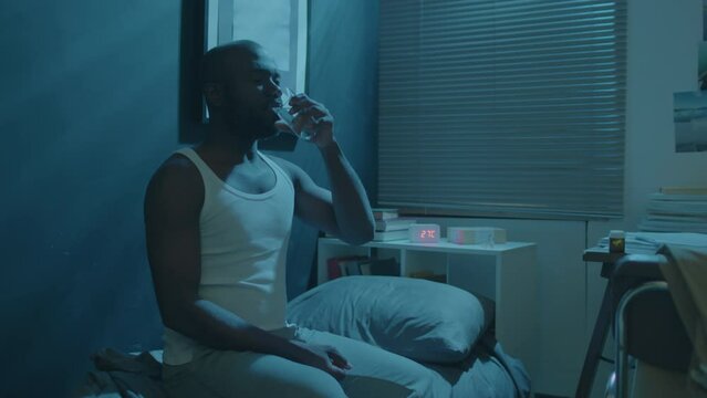 Black man sitting on bed in dark room, taking sleeping pills and drinking water while having insomnia at night