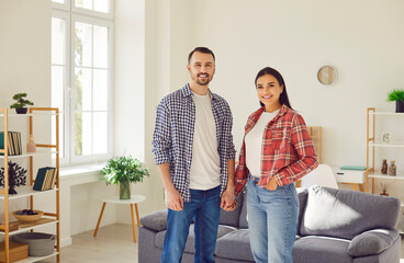 Portrait of happy loving family couple at home. Young man and woman in jeans and plaid and checkered shirts standing together and holding hands in modern living room interior with couch in background