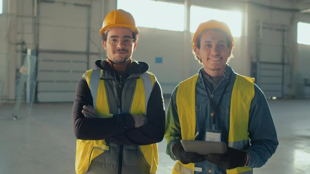 Medium portrait of two male Biracial construction workers in hardhats and protective workwear smiling and posing for camera with tablet in hands during workday inside empty spacious building