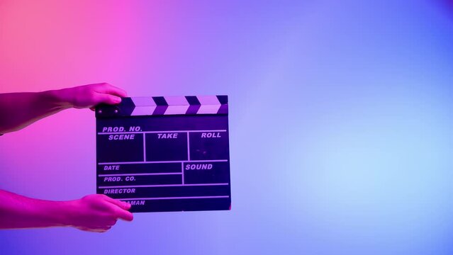 Hand is holding Black clapper board or movie slate on a blue background with a circular light, illuminated with pink neon light. Cinema industry, entertainment.