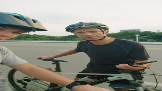 Vertical POV shot of boy and his dad in cycling helmets and riding clothing looking at camera and talking while filming vlog about bike trip