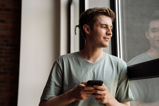 Portrait of smiling man using mobile phone while standing by the window