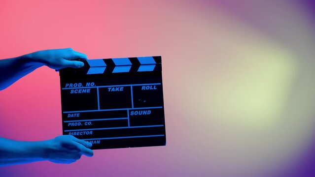 Hand is holding Black clapper board or movie slate on a pink background with a circular light, illuminated with blue neon light. Cinema industry, entertainment.