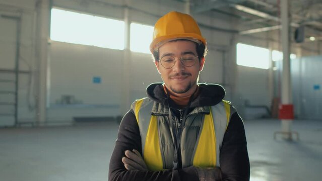 Chest up portrait of young smiling Biracial builder in glasses and safety hardhat posing for camera with folded arms during workday in empty indoor construction site on sunny day