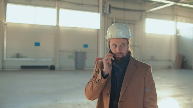 Tracking footage of Caucasian bearded building owner wearing white safety hardhat, suit and beige coat and speaking on smartphone while walking at indoor construction site