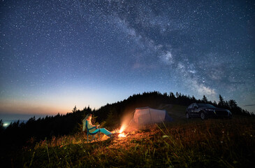 Night camping in the mountains under starry sky full of stars. Travelling woman admiring starry night in mountains. Young woman sitting next to camp fire, tourist tent and off-road car.