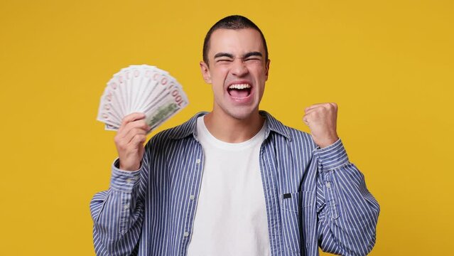 Happy young middle eastern man he wear blue shirt white t-shirt hold in hand fan of cash money in dollar banknotes doing winner gesture celebrate isolated on plain yellow background studio portrait