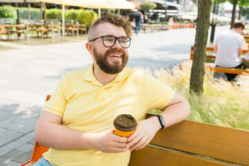 Millennial smiling student man in yellow polo t-shirt eyeglasses sitting on bench drink coffee have...