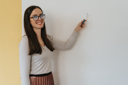 Brunette woman smiling and pointing to an empty wall with a marker.