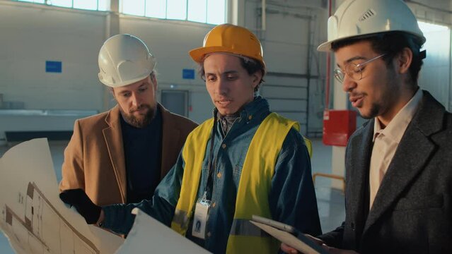 Medium side shot of smiling diverse business associates and engineer in hard hats discussing building plan execution at construction siteMedium side shot of smiling diverse business associates and eng