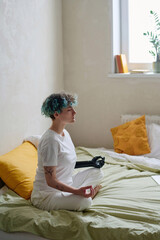 Young woman with green curly hair meditating on bed in the morning