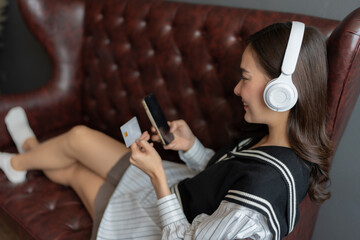 Close-up photo of a young Asian woman Wear headphones and listen to music. Sitting on the sofa, relaxing on a happy holiday. Looking at a smartphone and shopping, placing an order with a credit card